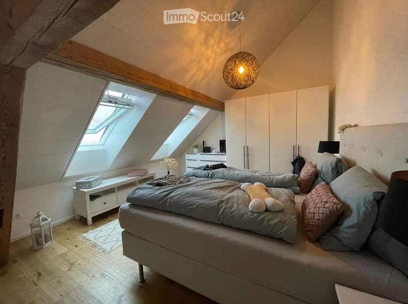 Cozy attic bedroom with wooden floors, slanted ceilings, large skylights, a double bed with pillows and blankets, a nightstand, storage cabinet, and decorative items.