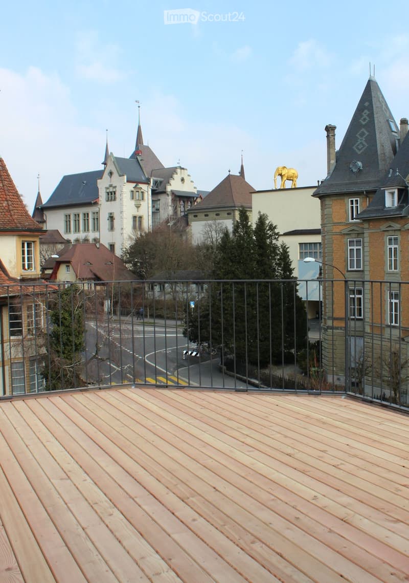 Wooden deck with metal railing overlooking a street lined with historic buildings and trees.