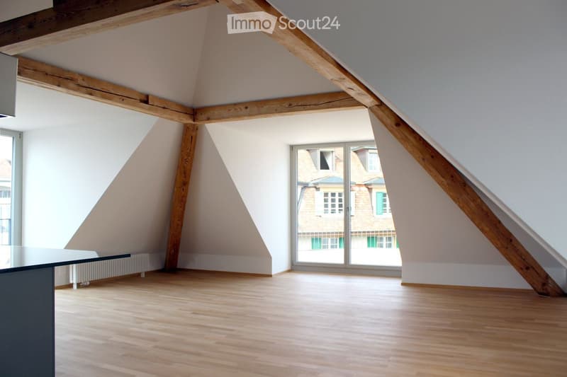 An empty attic room with wooden beams, wooden flooring, and large windows offering a view of the outside. The room has white walls and a modern, minimalist design.