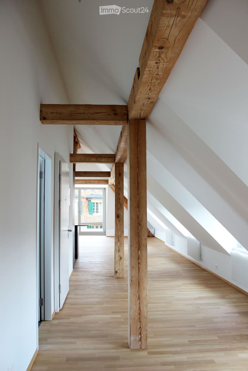 Bright, airy attic corridor with wooden beams and light wooden flooring, leading to a room with large windows and a view of the outside.
