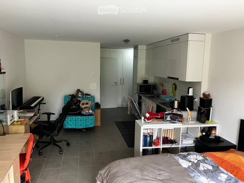A multi-functional room combining a bedroom and a kitchen area. Features a bed with a patterned blanket, a desk with a chair, a guitar on a couch, a piano, and a kitchen area with cabinets, a refrigerator, and a microwave.