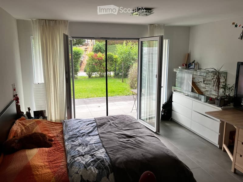 Bright bedroom with a large bed, nightstand, lamp, and a sliding glass door leading to a garden. There is a small table, cabinet, and a plant in the corner.