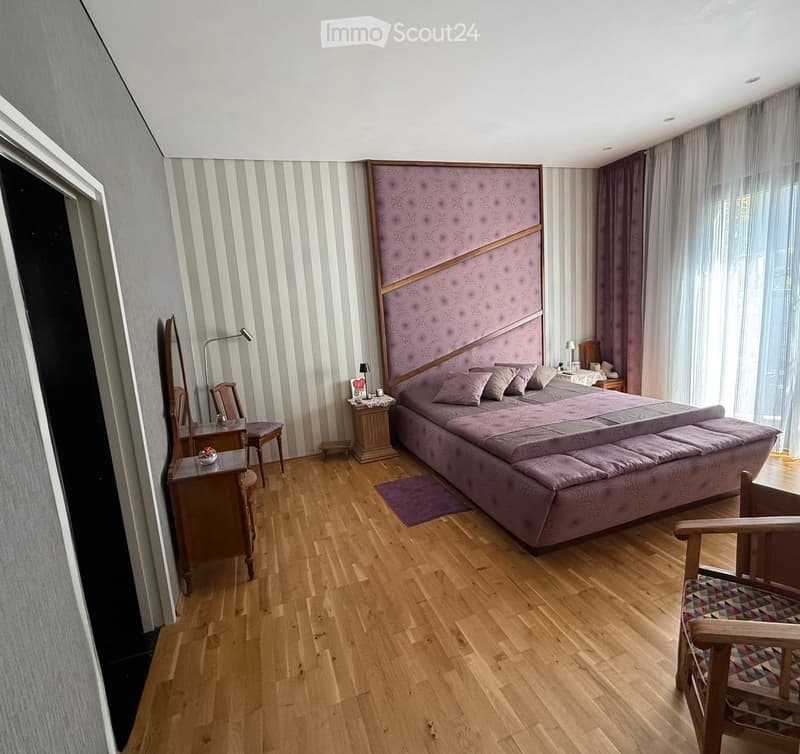 large bed with pillows, purple headboard, wooden floor, open window with white curtains, bedside table with lamp, mirror, wooden chair, wooden desk with lamp, small table, various decorative items