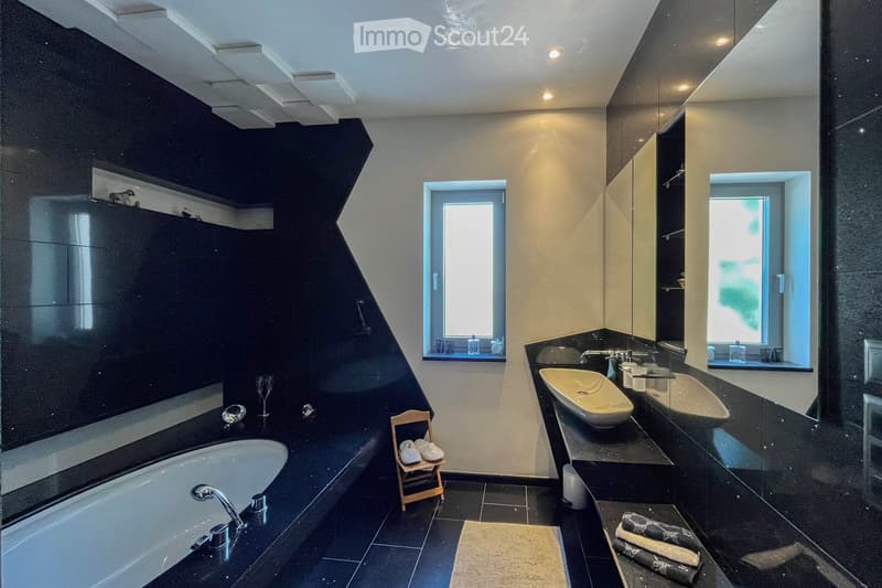 Modern bathroom with black tiles, a bathtub, two sinks, a small wooden stool with towels, wine glasses, and a mirror. The room features large windows and sleek, contemporary design elements.