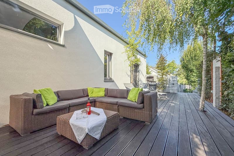 Outdoor wooden deck, brown wicker L-shaped sofa, green cushions, center table with white cloth, bottles, glasses, wooden flooring, white wall, clear blue sky, greenery, trees