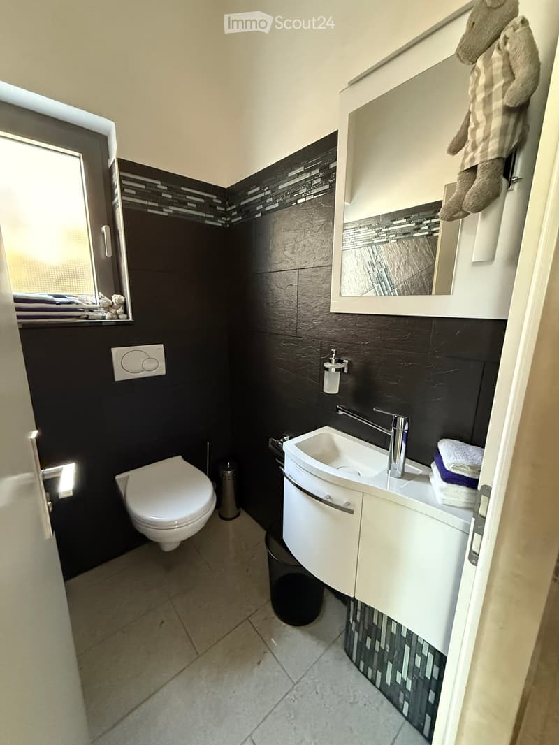 White toilet, silver sink with faucet, black tile walls, mirror, window with towels, small trash bin