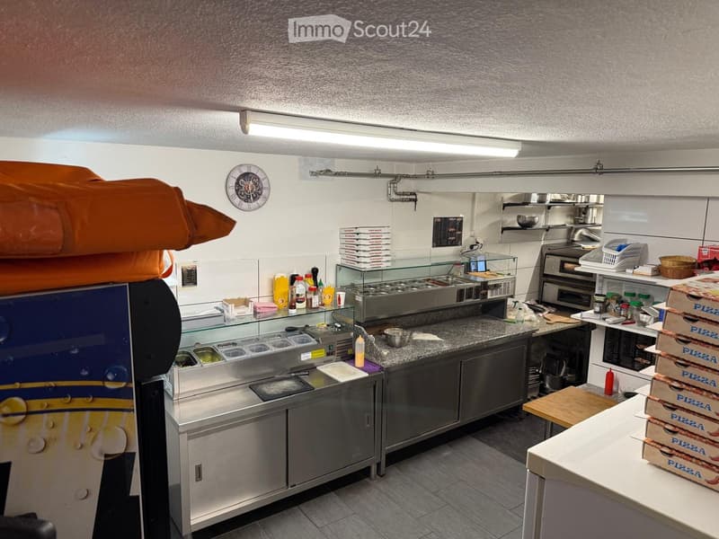 Commercial kitchen with stainless steel appliances, granite countertop, display case, and pizza boxes stacked on a table.