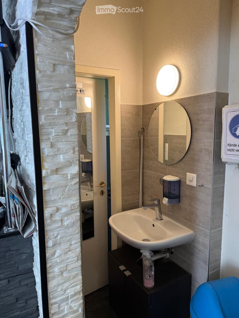 Modern bathroom with a white sink, round mirror, and a small cabinet holding a bottle of hand sanitizer. The walls are tiled in a light gray color, and there is a round light fixture above the mirror.