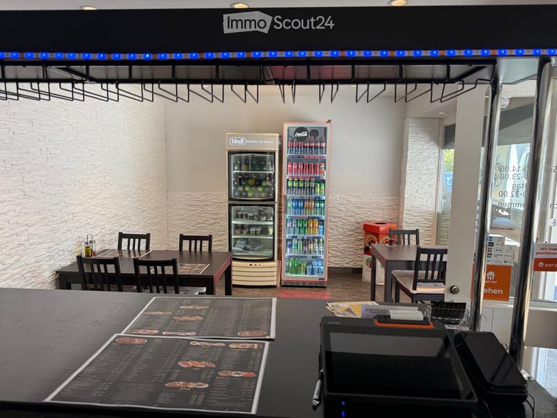 Restaurant interior with tables, chairs, refrigerators, and a cash register. Menu boards are placed on the counter.
