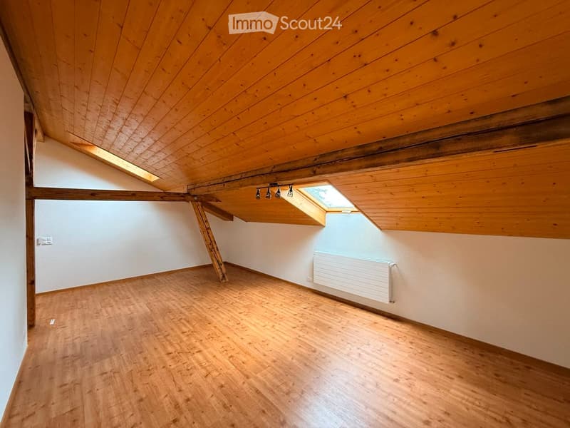 Empty attic room with wooden ceiling, wooden floor, white walls, skylight windows, and a radiator on one wall.