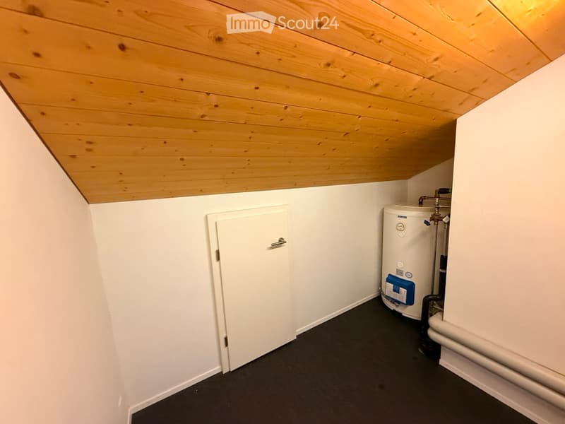 An unfurnished utility room with a wooden slanted ceiling, white walls, black flooring, a white door, and a water heater with pipes.