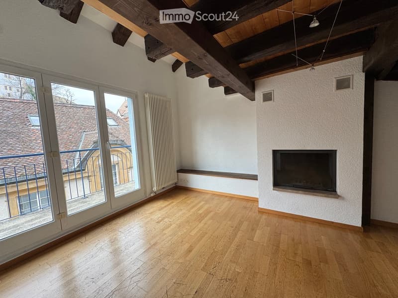 Empty room with wooden flooring, exposed wooden beams, fireplace, large windows with balcony access, radiator, and visible ventilation grilles.
