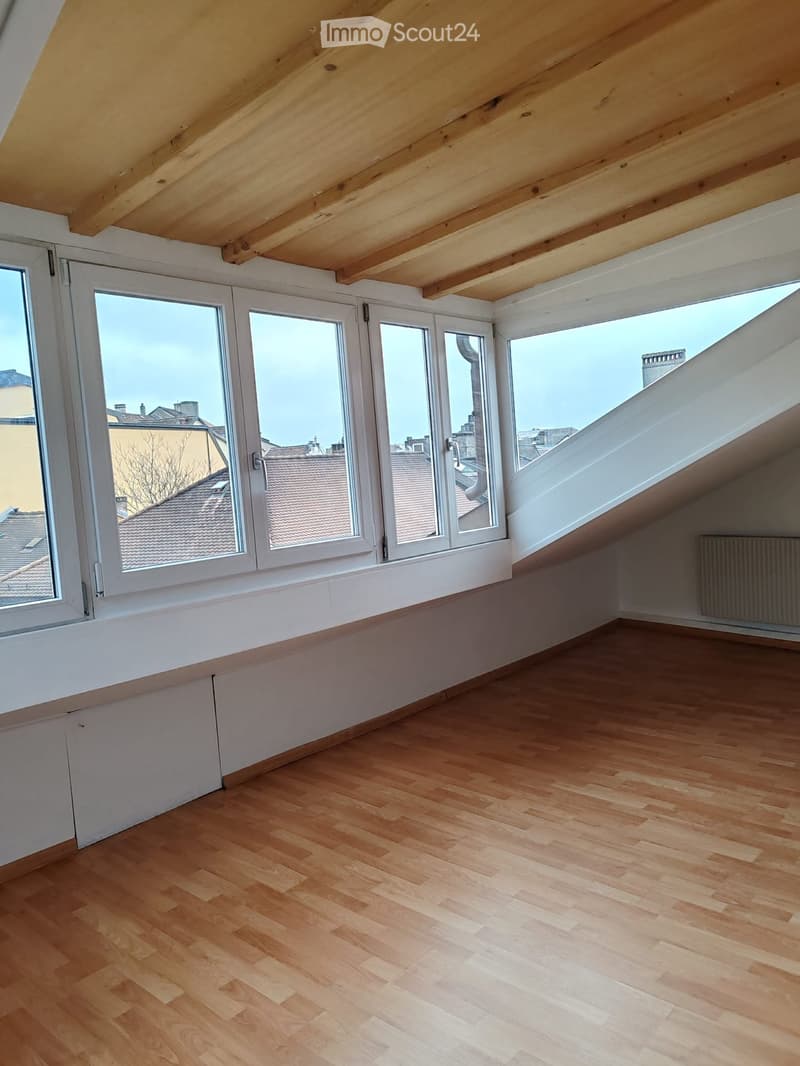 Empty attic space with wooden flooring, large windows offering a view of rooftops, and exposed wooden beams on the ceiling.