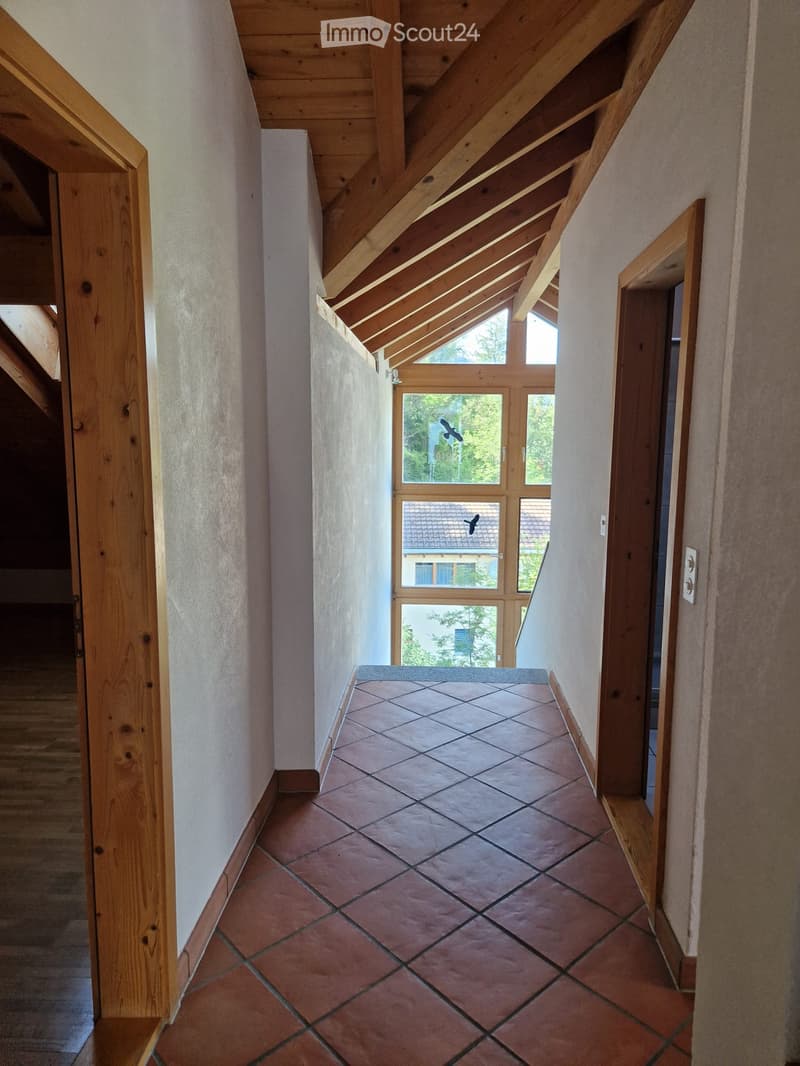 A narrow hallway with exposed wooden beams on the ceiling, tiled floor, and large windows at the end showing a view of the outdoors.