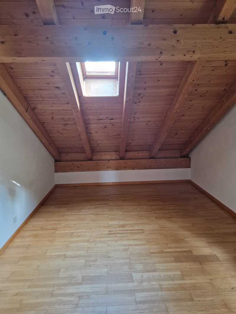 Empty room with wooden floor and exposed wooden ceiling beams, featuring a small skylight window.