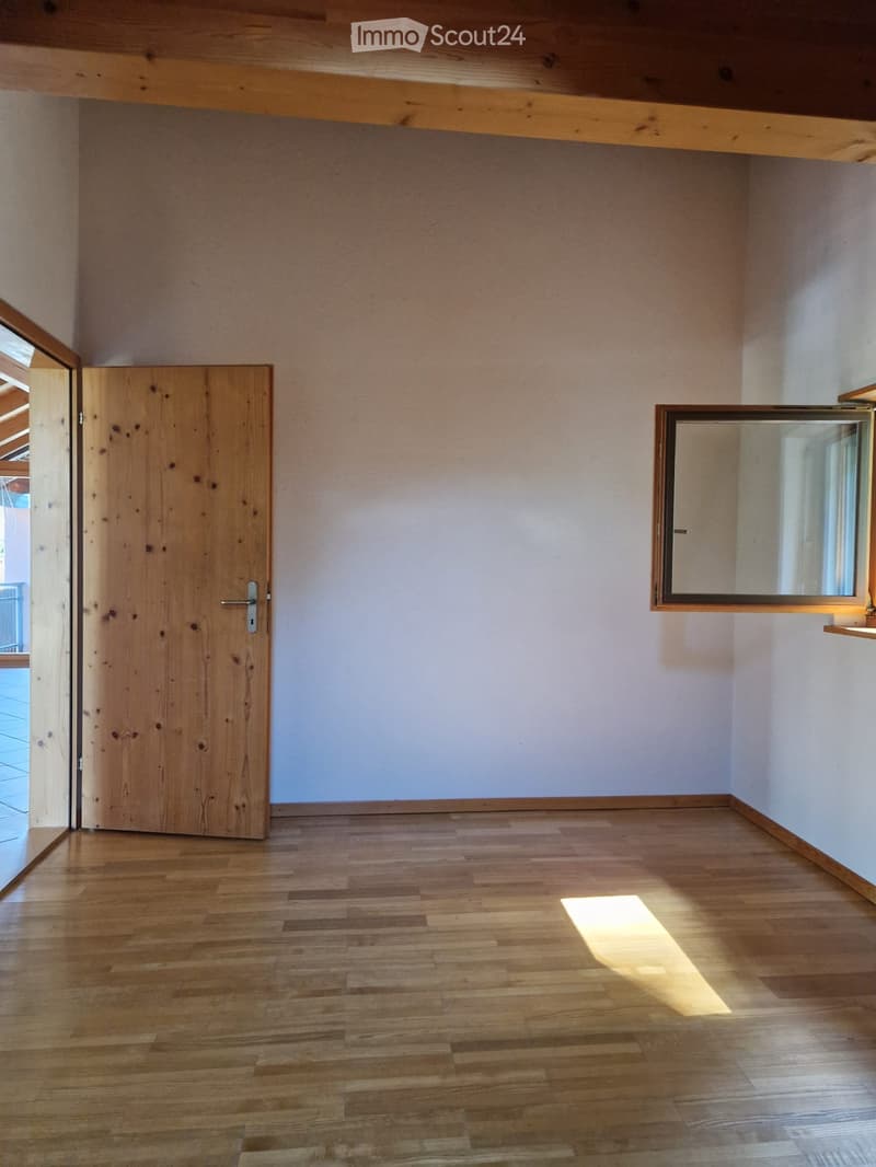 Empty room with wooden floor, light-colored walls, and a wooden door leading to another space. A small window with a mirror is mounted on the wall.