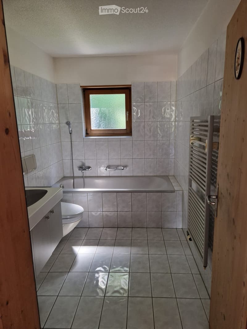 Modern bathroom with grey tiles, a bathtub, toilet, sink, and a radiator. Features a frosted window and a wooden door frame.