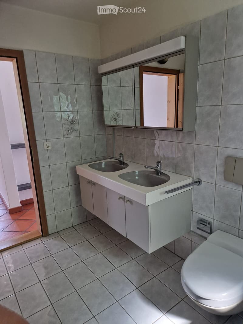 Modern bathroom with double sink, mirror, and toilet. Grey tiled walls and floor, white cabinetry, and wooden door frame.