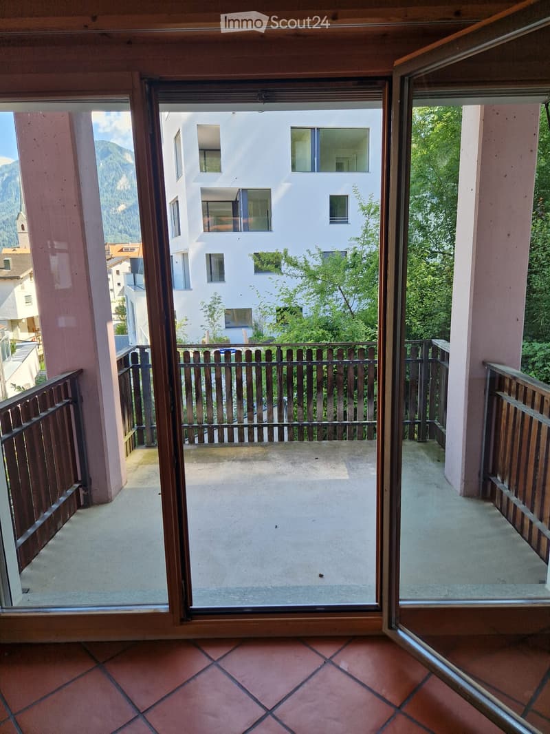 Balcony with wooden railings, overlooking a building and green trees, with a scenic mountain view in the background.