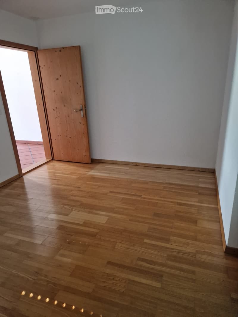 Empty room with wooden flooring and light-colored walls, featuring an open wooden door leading to another space.