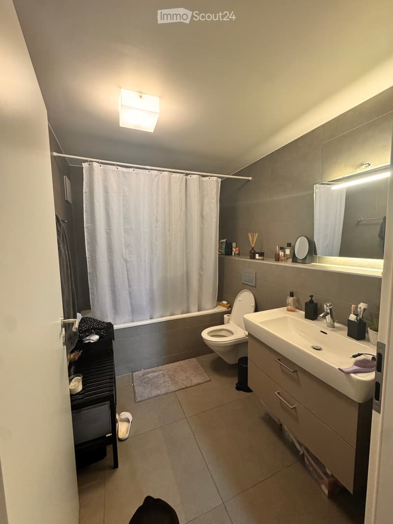 Modern bathroom with a white sink, toilet, bathtub, and large mirror. Includes a shower curtain, a small cabinet, and a towel on the floor.