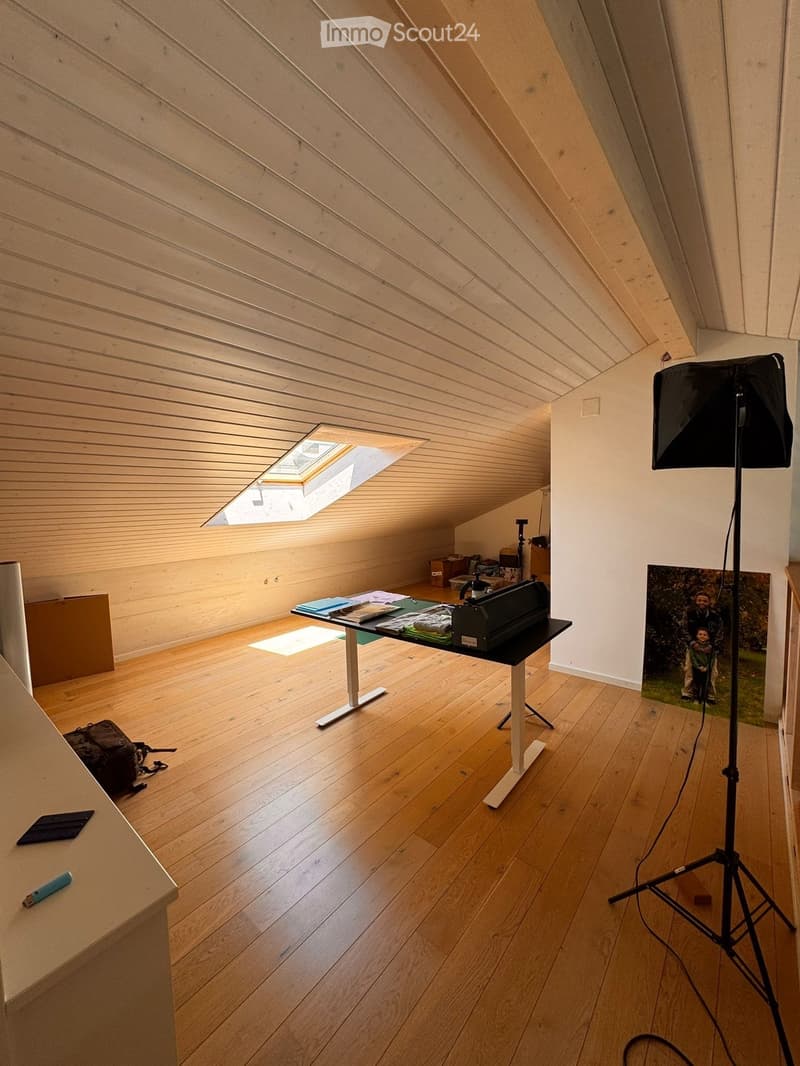 An attic room with wooden floors and slanted wooden ceilings, featuring a skylight window, a small table with various items, a camera tripod, and a picture frame on the wall.