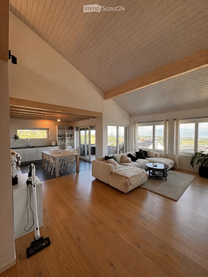 Open-plan living and dining area with wooden flooring, large windows offering a scenic view, a sectional sofa, coffee table, dining table with chairs, bookshelf, and kitchen area.