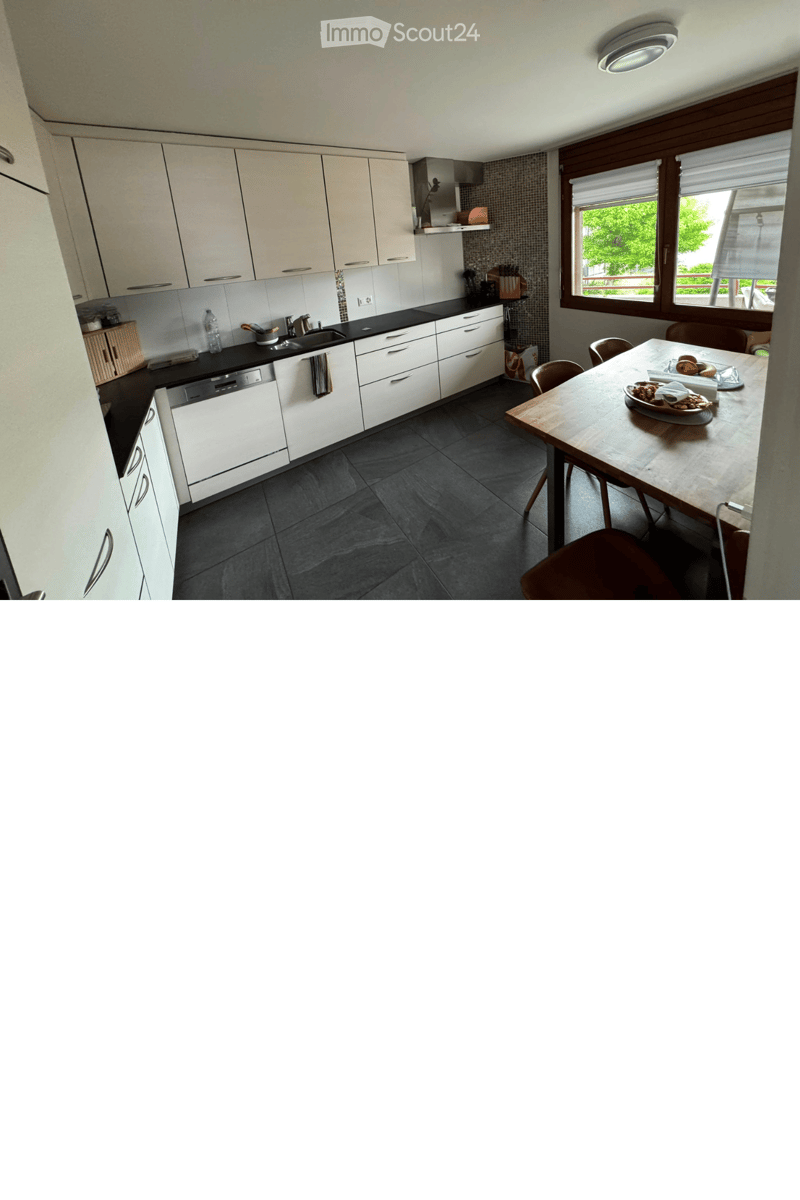 Modern kitchen with white cabinets, black countertops, and a dining area with a wooden table and chairs. Features a sink, dishwasher, and various kitchen items on the counter.