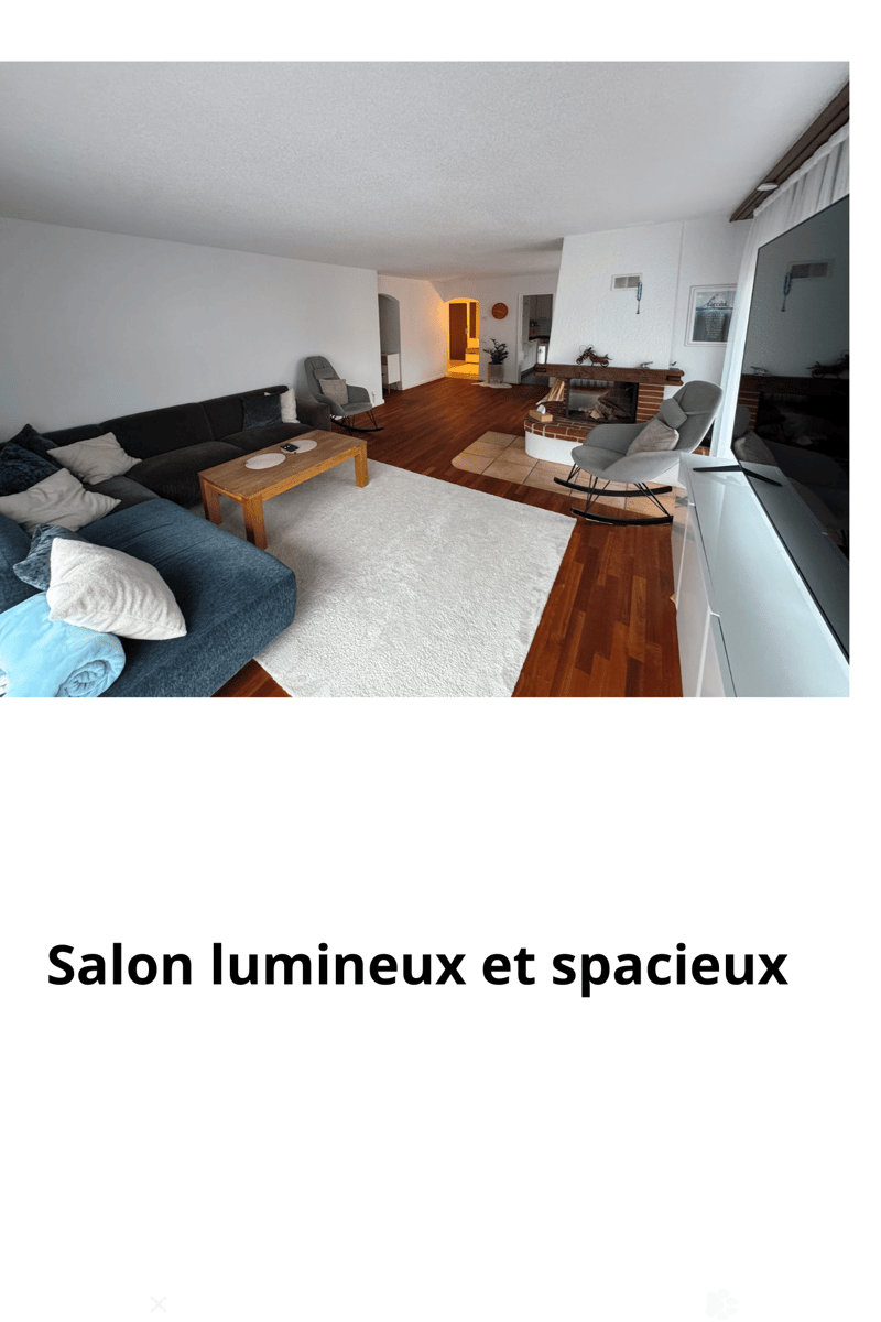 Bright and spacious living room with a dark blue sectional sofa, wooden coffee table, armchairs, fireplace, and television. The room features hardwood flooring and large windows allowing natural light to fill the space.
