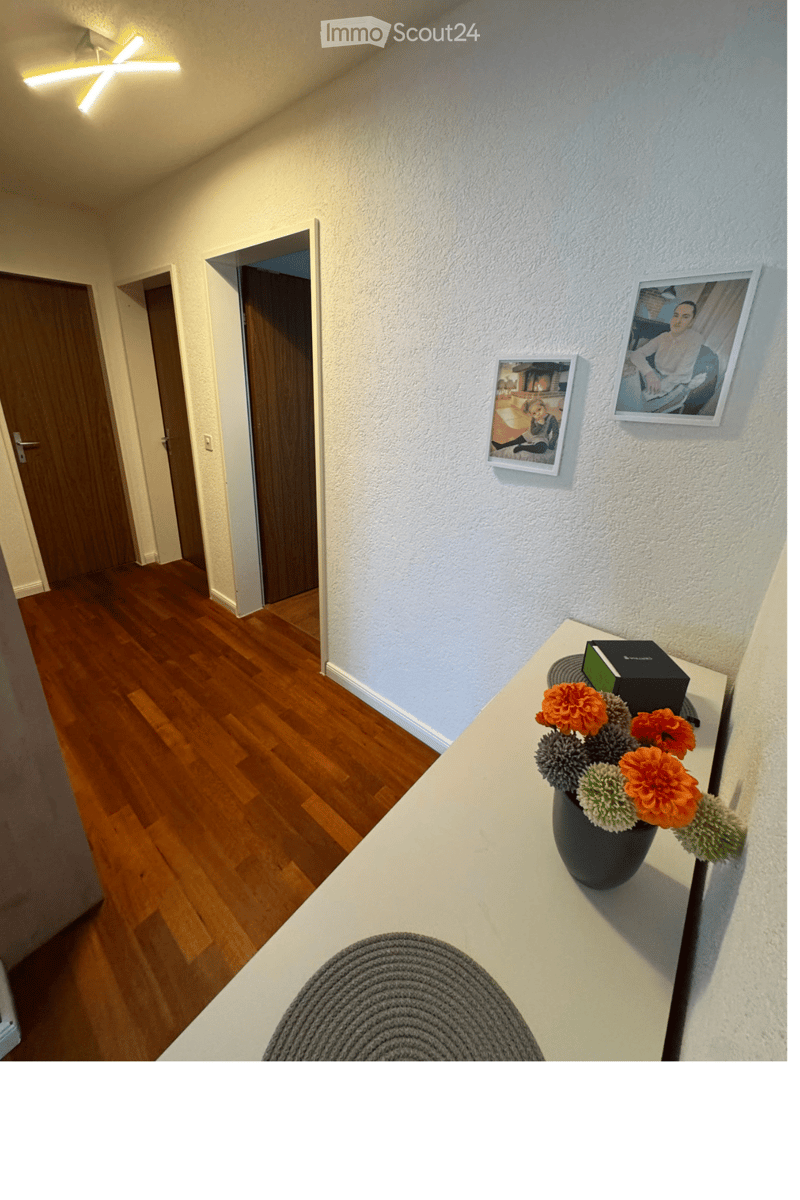 Hallway with wooden flooring, white walls, and framed pictures. A small table with a vase of flowers and a grey rug is visible.