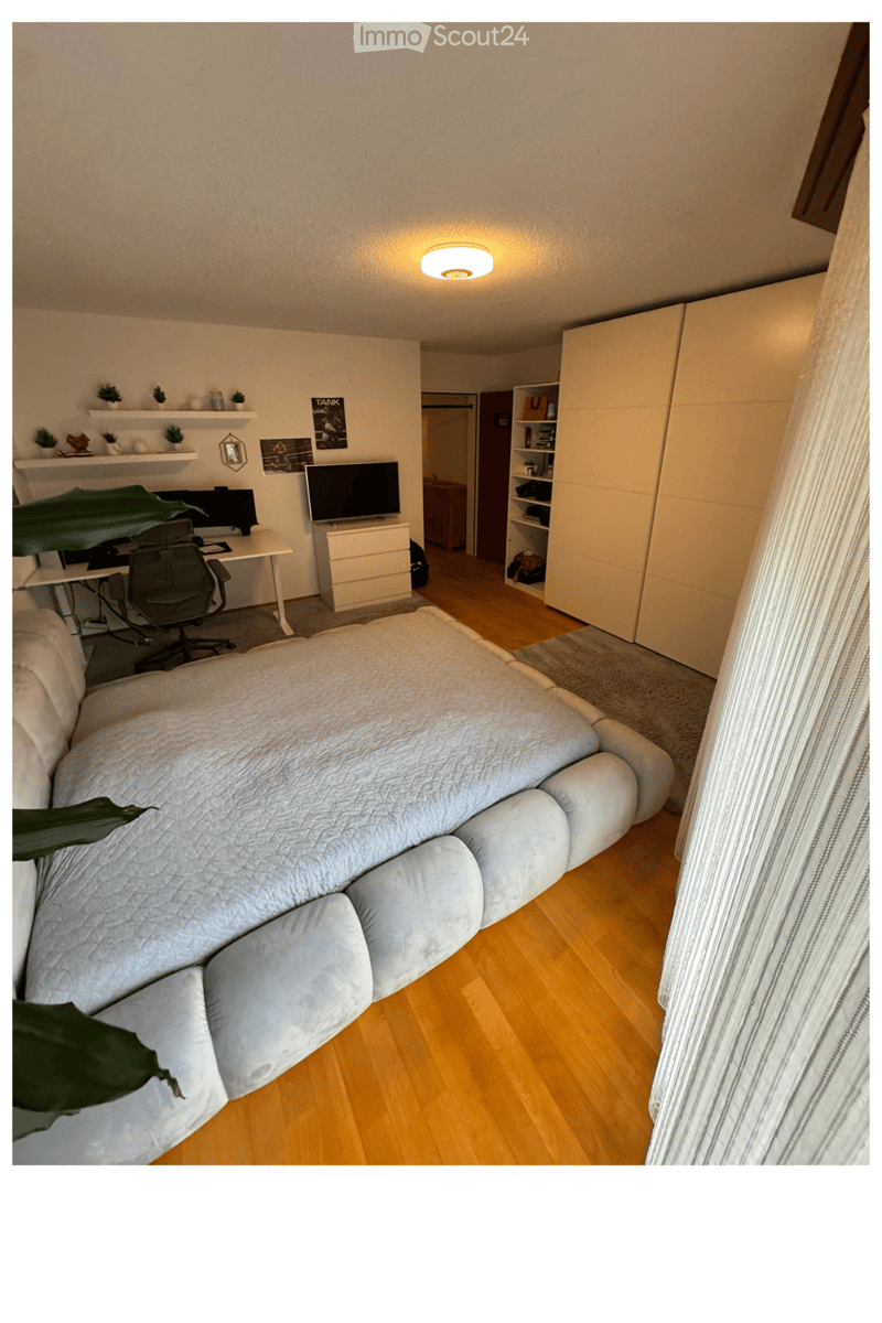 A well-lit room with a sofa, desk, chair, TV, and shelves. The room features wooden flooring and a large window with a curtain.