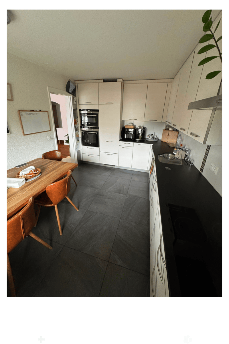 Modern kitchen with white cabinets, black countertops, and a wooden dining table with chairs. Appliances include an oven, coffee maker, and sink.