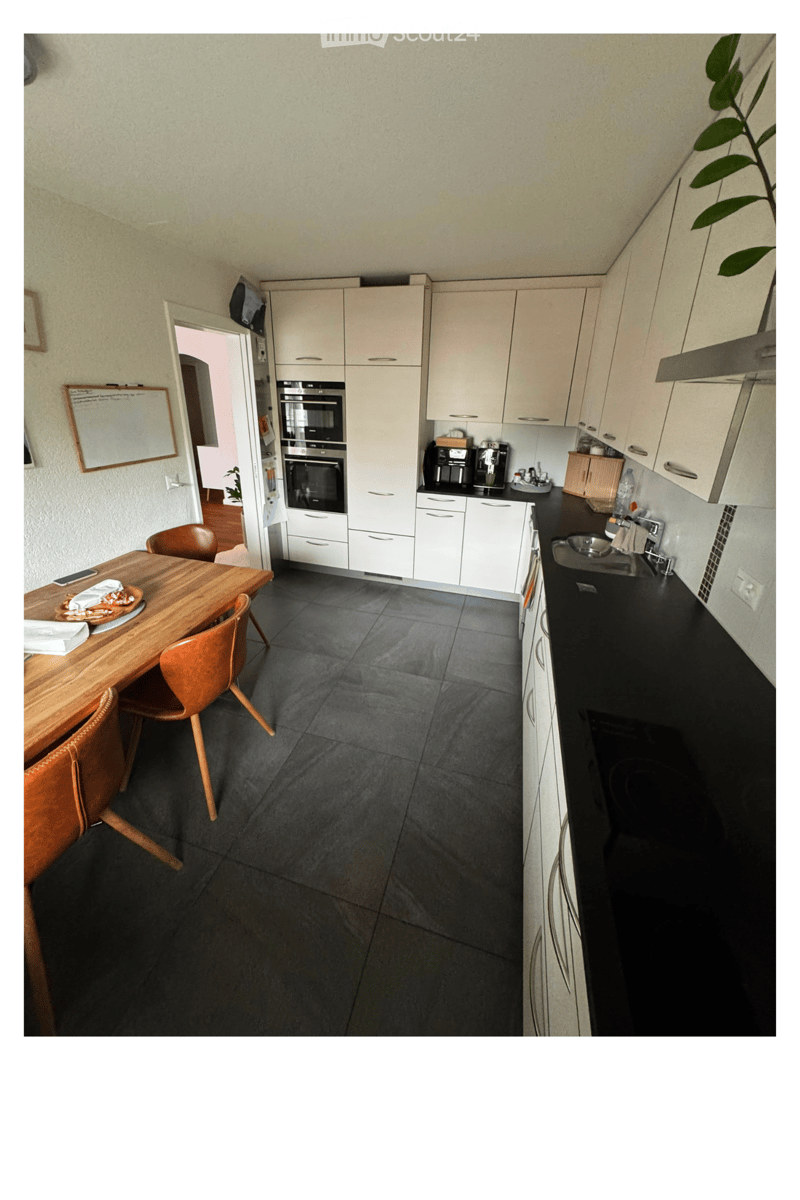 Modern kitchen with white cabinets, black countertops, and a wooden dining table with chairs. Appliances include an oven, coffee maker, and sink.