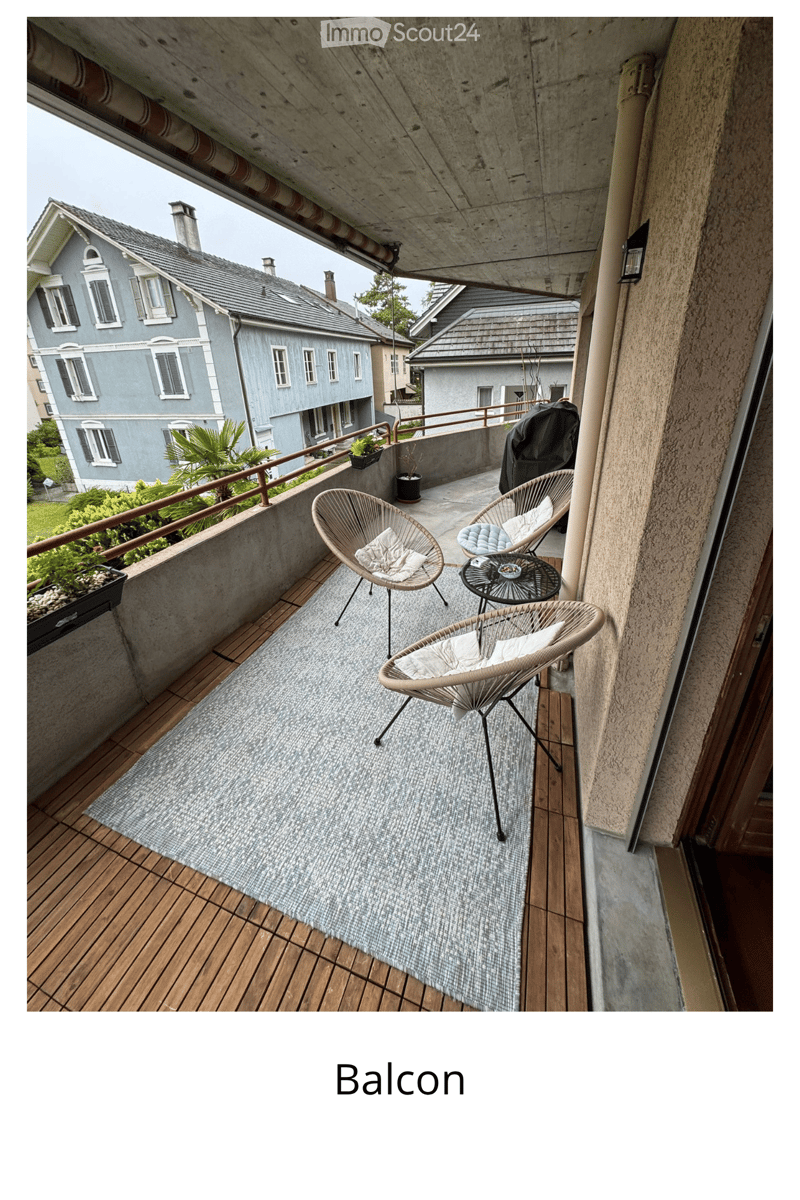 Balcony with wooden decking, wicker chairs, small table, and potted plants, overlooking a residential area with multi-story buildings.