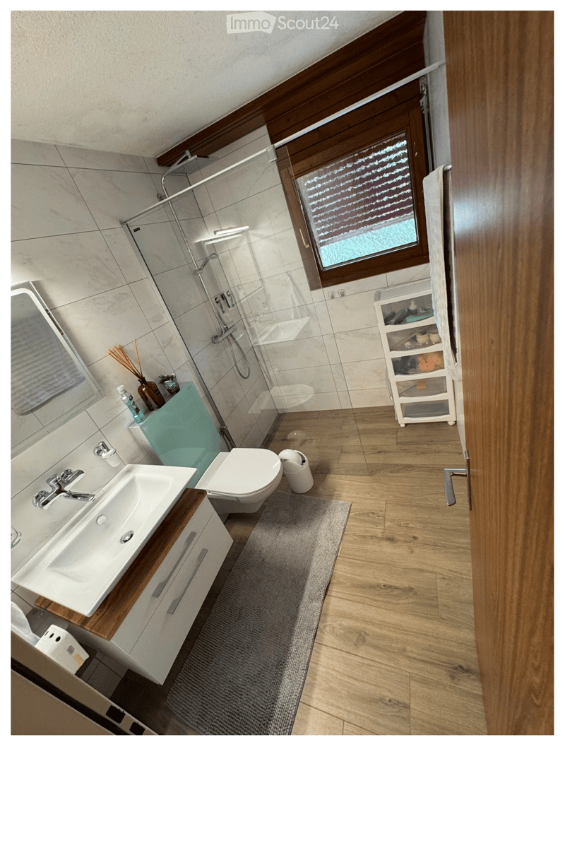 Modern bathroom with a white sink, toilet, glass-enclosed shower, and a wooden vanity. Features a grey rug, a small storage cabinet, and a window with frosted glass.