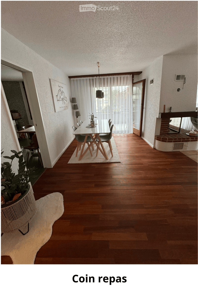 Dining room with wooden flooring, a modern dining table with chairs, a potted plant, a mirror, and artwork on the wall. The room features large windows with sheer curtains and a view of the outdoors.