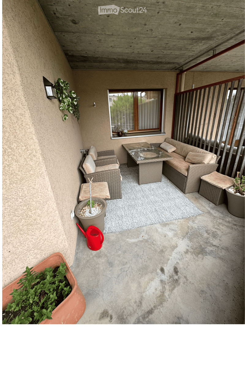 Balcony with concrete floor, wicker furniture including a sofa, table, chairs, and a stool, along with potted plants and a watering can.