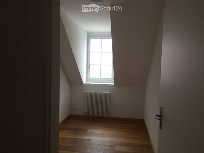 Empty attic room with wooden flooring, white walls, a single window, and a radiator. No furniture or equipment present.