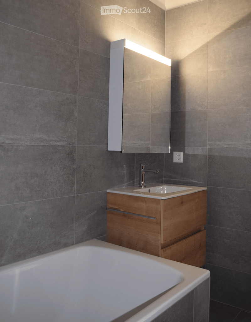 Modern bathroom with a white bathtub, wooden sink cabinet, sleek faucet, and illuminated mirror above the sink.