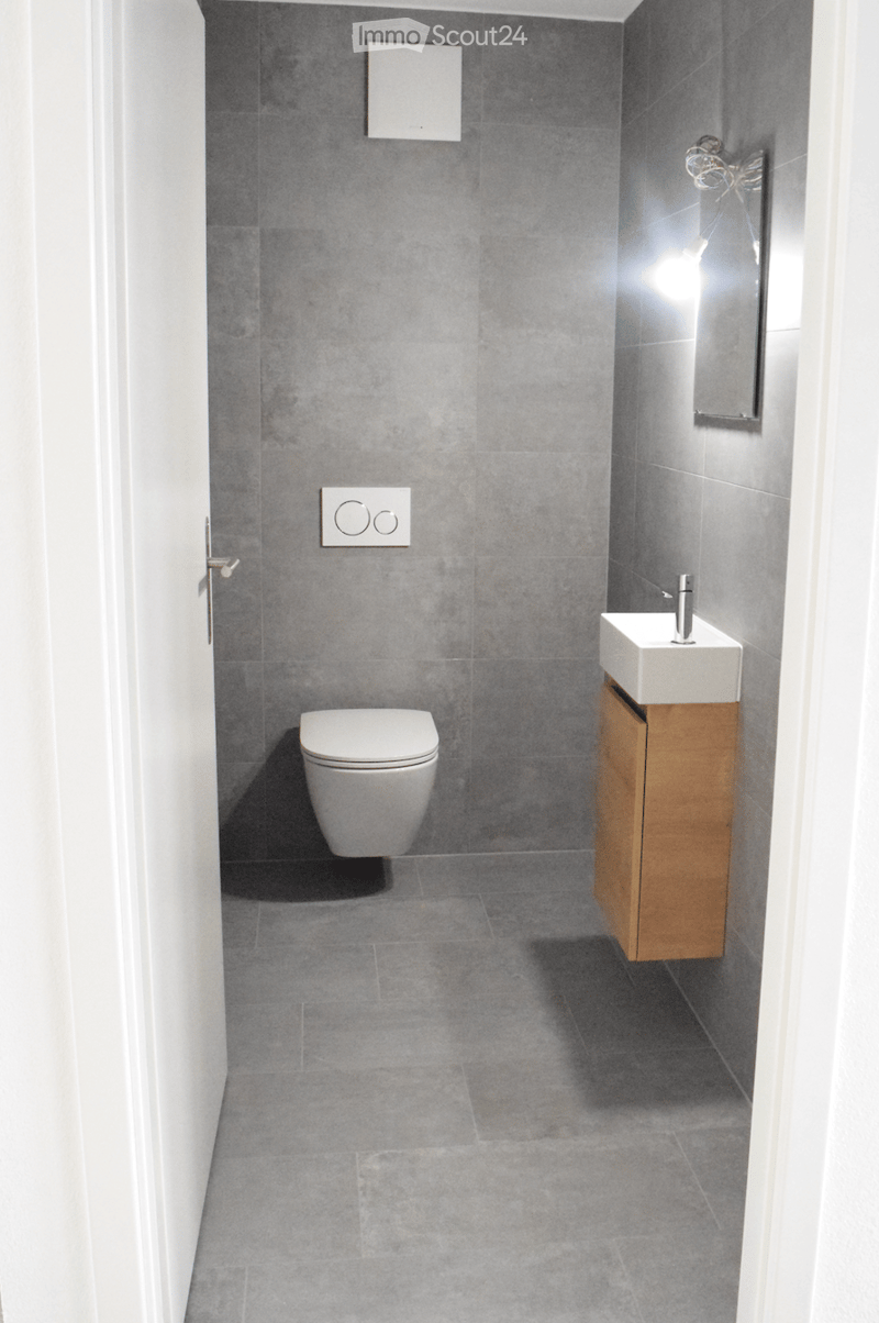Modern bathroom with grey tiled walls and floor, white toilet, white sink with wooden cabinet, and a mirror above the sink.