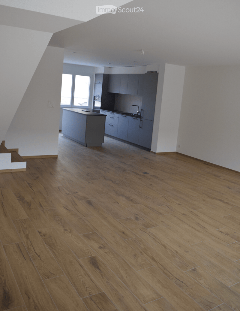 Open-plan space with wooden flooring, white walls, and a modern kitchen area featuring cabinets and a countertop. The room is completely empty with no furniture or personal items.