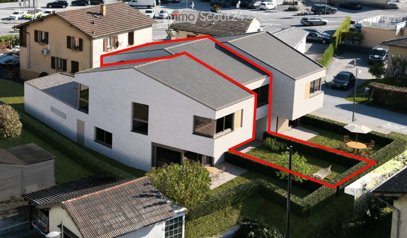 Aerial view of a modern two-story house with a grey roof, large windows, and a garage. The house is surrounded by a well-maintained garden with outdoor seating and a parking area.