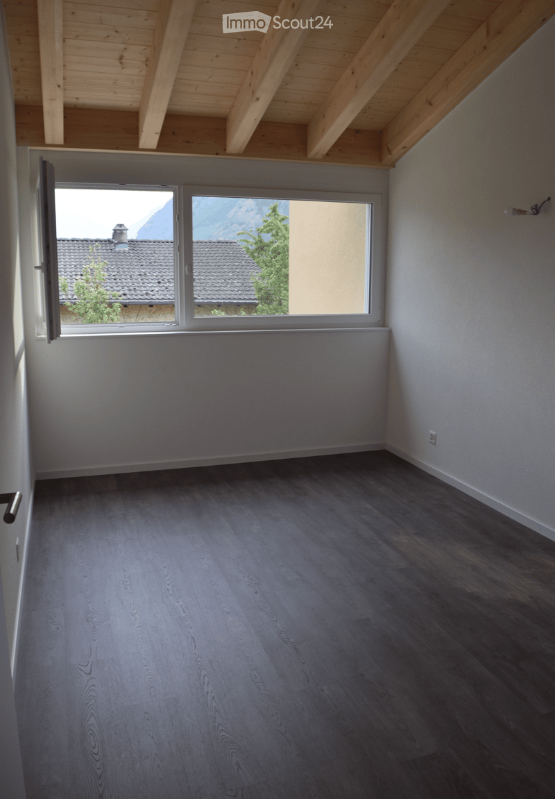 Empty room with dark wooden flooring, light grey walls, and large windows offering a view of a roof and mountains.