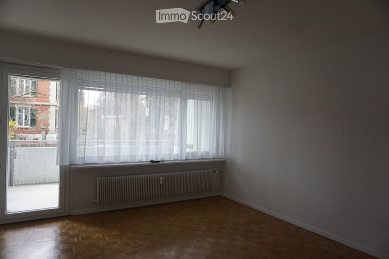 Unfurnished Room. Empty room with wooden flooring, large window with white curtains, radiator, and a sliding glass door leading to a balcony.