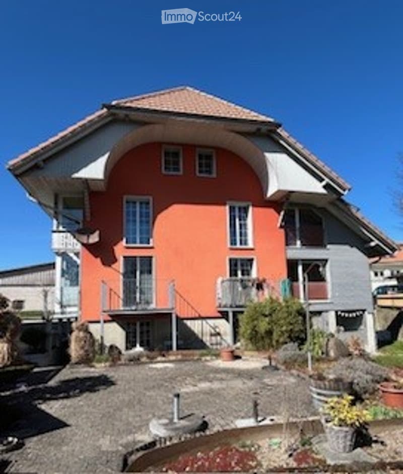 Modern two-story house with orange facade, white trim, multiple windows, balconies, and a small garden area with potted plants.