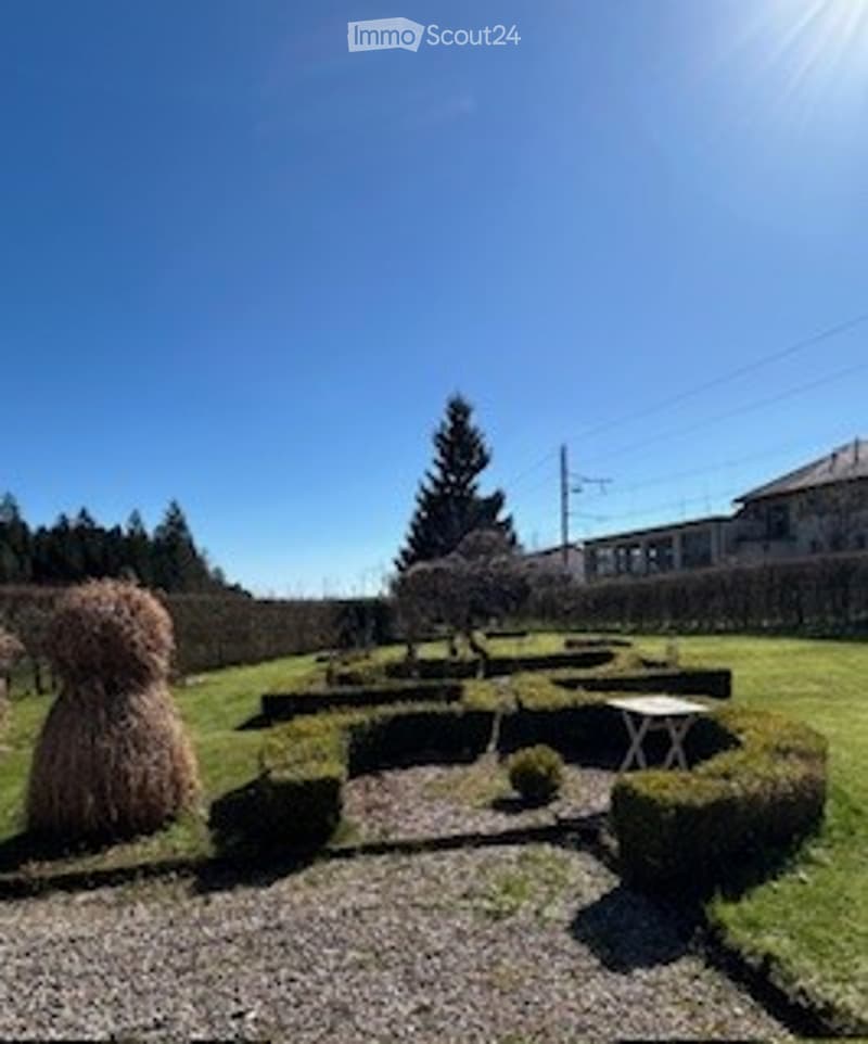 Well-maintained garden with neatly trimmed hedges, gravel pathways, a small table, and a large straw bale sculpture.