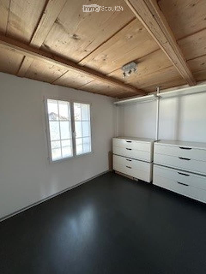 Empty room with white walls, dark flooring, large window, and built-in white storage units.