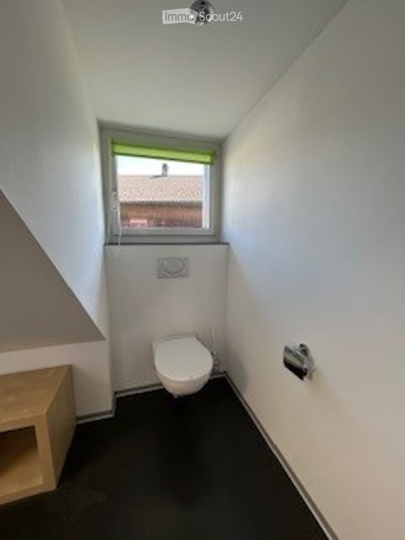 Small bathroom with a toilet, window, and light fixture. The walls are white, and the floor is dark.