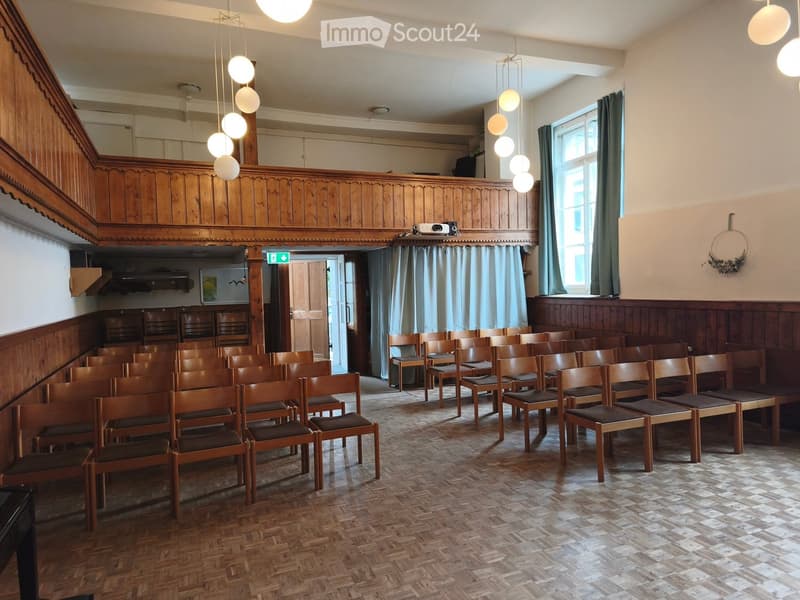 Wooden chairs arranged in rows, wooden panel walls, large windows with curtains, projector on a shelf, decorative wreath on wall.