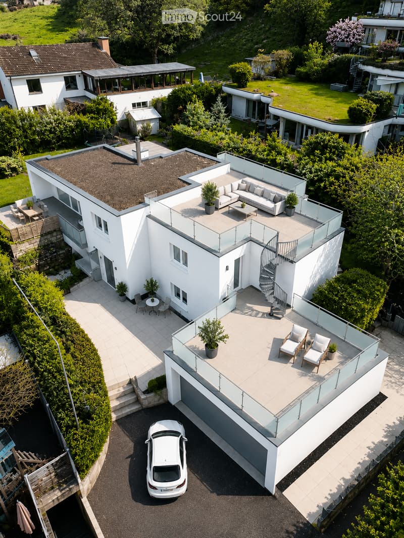 Aerial view of a modern white house with multiple levels, featuring a rooftop terrace with glass railings, outdoor seating, and a parked car in the driveway.
